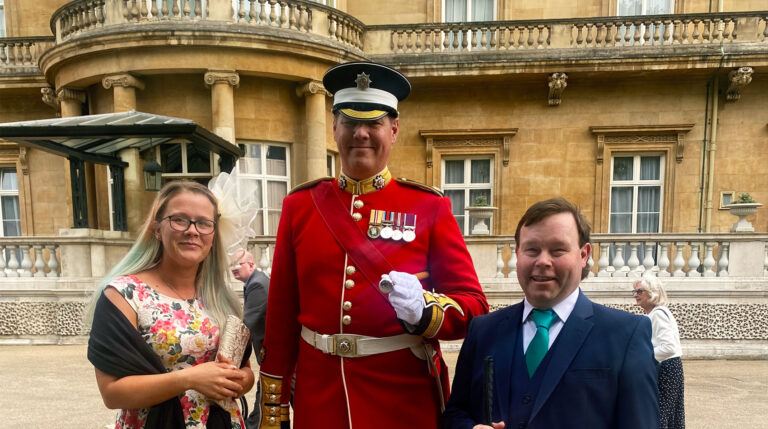 Representatives-Martyn-and-Stacey-smiling-with-a-royal-guard-outside-Buckingham-Palace